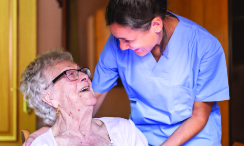 A healthcare worker in blue scrubs embraces and smiles warmly at an elderly woman with glasses and curly gray hair who is seated and looking up at her with a joyful expression.