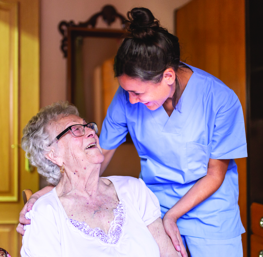 A healthcare worker in blue scrubs embraces and smiles warmly at an elderly woman with glasses and curly gray hair who is seated and looking up at her with a joyful expression.