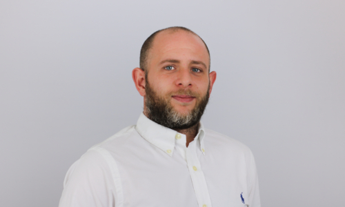 A man with a beard wearing a white button-down shirt with a small logo stands against a light gray background in what appears to be a professional headshot or portrait photo.