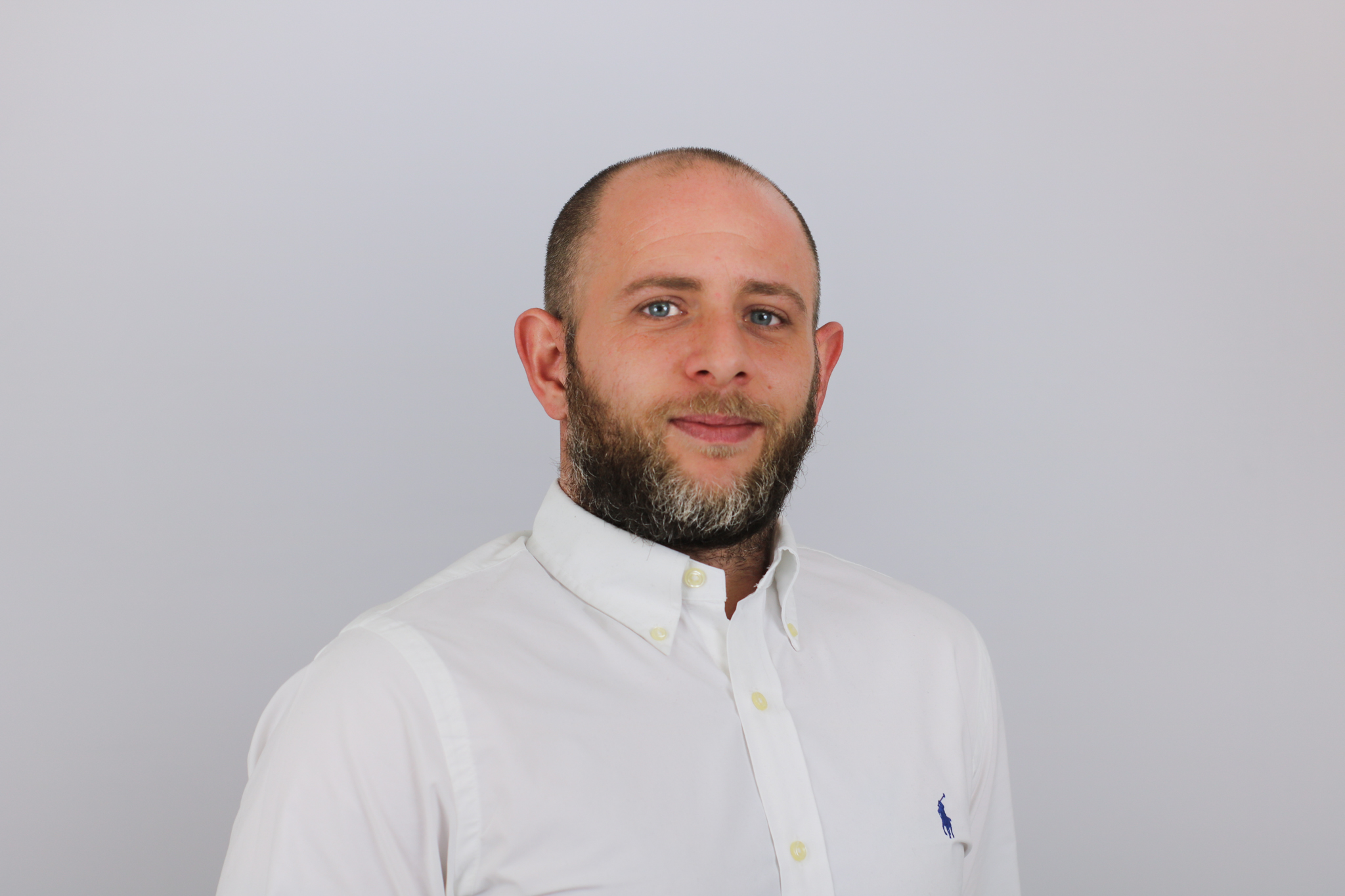 A man with a beard wearing a white button-down shirt with a small logo stands against a light gray background in what appears to be a professional headshot or portrait photo.