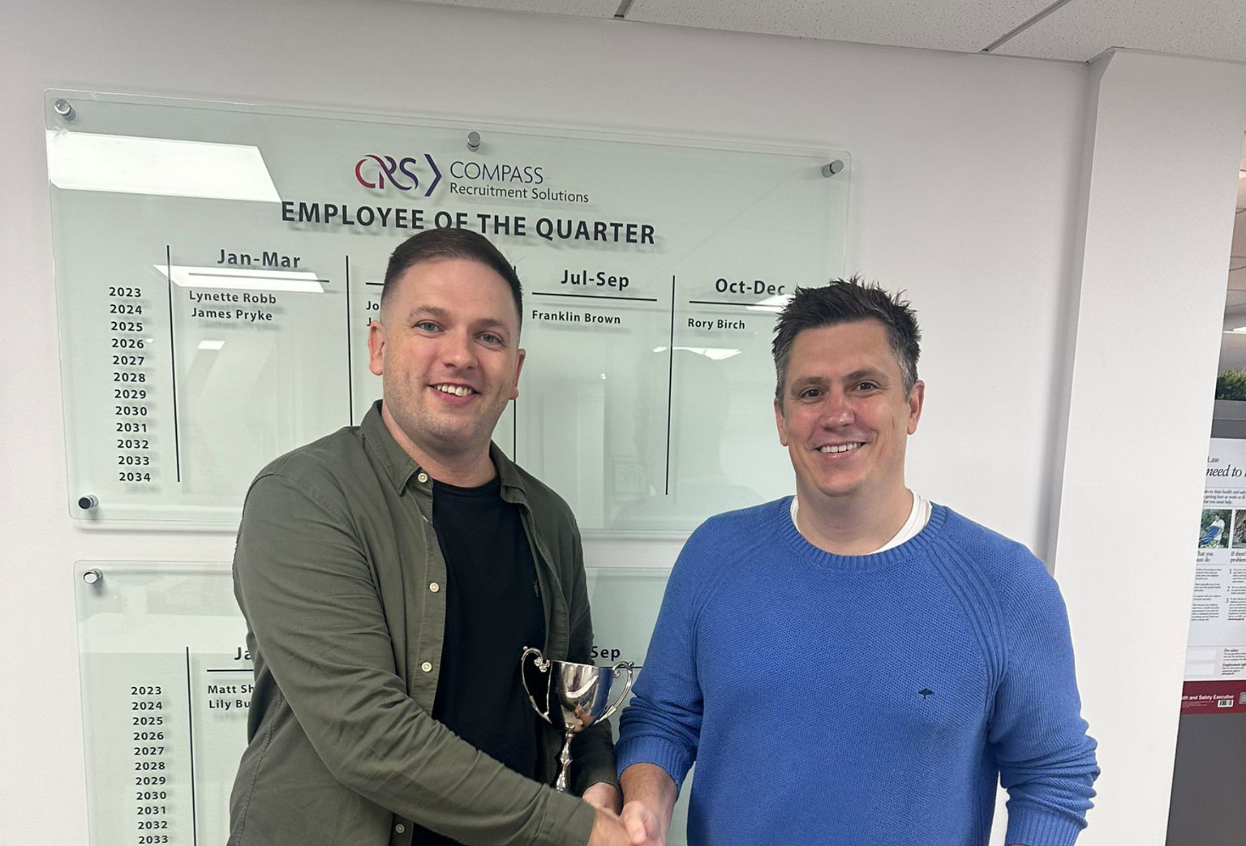 Two men in an office setting shake hands while holding a trophy, standing in front of an "Employee of the Quarter" board displaying names and years from a recruitment company called CRS Compass Recruitment Solutions.