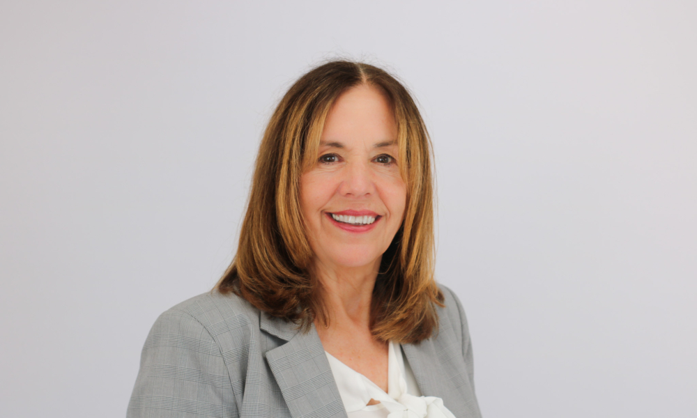 A professional woman with shoulder-length brown hair smiles warmly at the camera, wearing a light gray blazer over a white blouse against a plain white background.