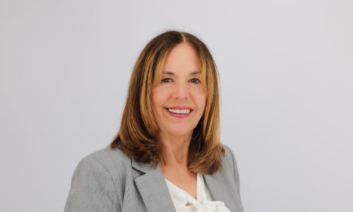 A professional woman with shoulder-length brown hair smiles warmly at the camera, wearing a light gray blazer over a white blouse against a plain white background.