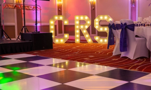 A formal event venue with a black and white checkered dance floor, illuminated marquee letters spelling "ORS" in the background, DJ equipment on the left, and white-covered chairs with navy blue sashes under colorful ambient lighting.