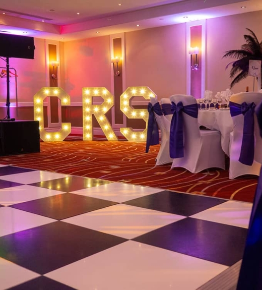 A wedding reception venue with a black and white checkered dance floor, illuminated "MRS" marquee letters, white chairs with navy blue sashes, and DJ equipment, all bathed in purple and pink ambient lighting.