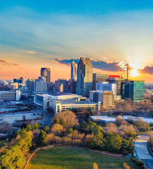 An aerial view of a mid-sized city skyline at sunset, featuring modern high-rise buildings and skyscrapers surrounded by green parkland and tree-lined streets. The dramatic sky displays vibrant orange and blue hues as the sun sets behind clouds, casting warm light across the urban landscape.