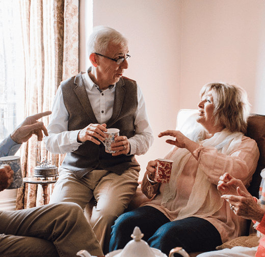 Four older adults sit in a bright, comfortable living room having an animated conversation, with two men and two women enjoying drinks together on a couch near a large window.