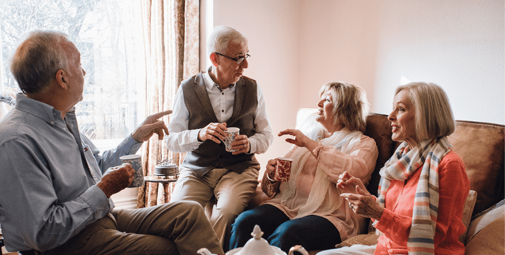 Four older adults sit in a bright, comfortable living room having an animated conversation, with two men and two women enjoying drinks together on a couch near a large window.