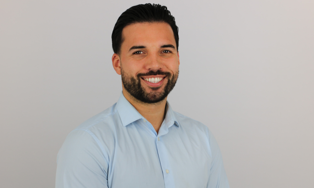 A smiling man with dark hair and a beard wearing a light blue dress shirt poses against a neutral background in what appears to be a professional headshot or portrait.