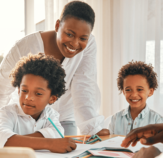 A smiling woman stands behind two young children who are sitting at a table with books and pencils, while a man sits across from them in what appears to be a bright, sunny learning environment.