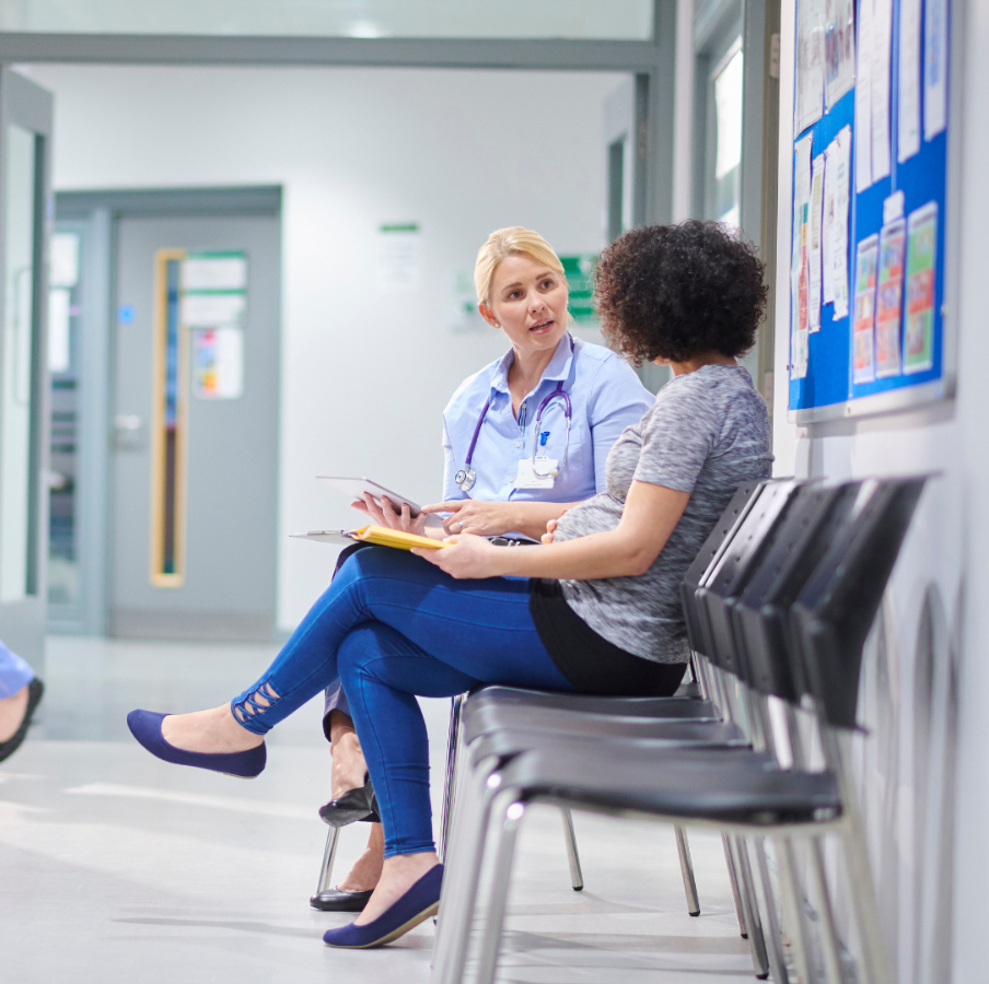 A healthcare professional with a stethoscope sits beside a young patient in a hospital waiting area, reviewing medical documents together while having a consultation.