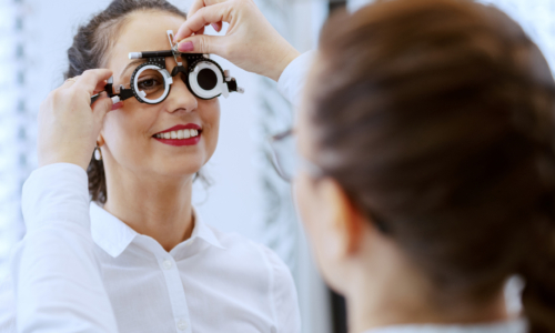 A smiling woman wearing a trial frame with multiple lens options during an eye examination, with an optometrist adjusting the device in a clinical setting with an eye chart visible in the background.
