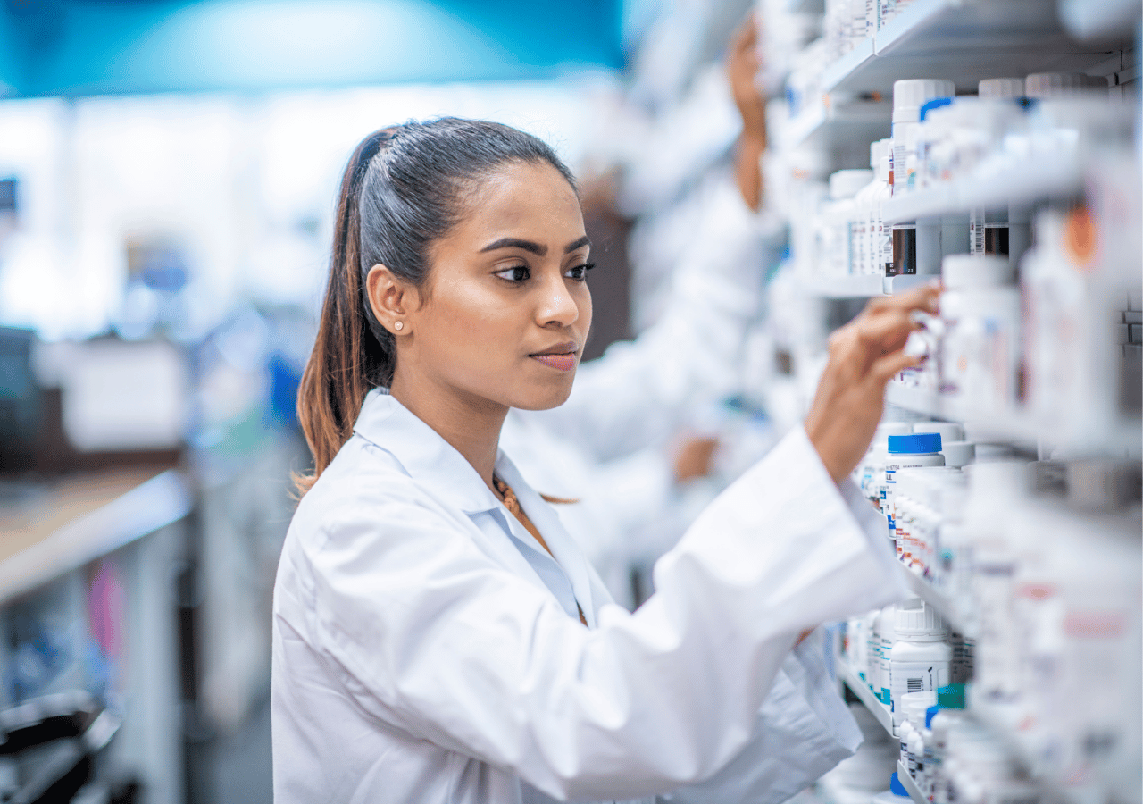 A female pharmacist in a white lab coat reaches for medication on shelves stocked with pharmaceutical products in a modern pharmacy.