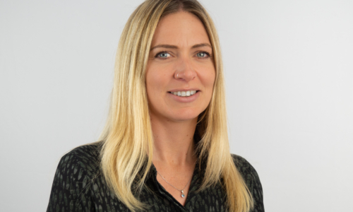 A professional headshot of a woman with shoulder-length blonde hair wearing a dark patterned blouse and a delicate necklace, smiling at the camera against a neutral gray background.