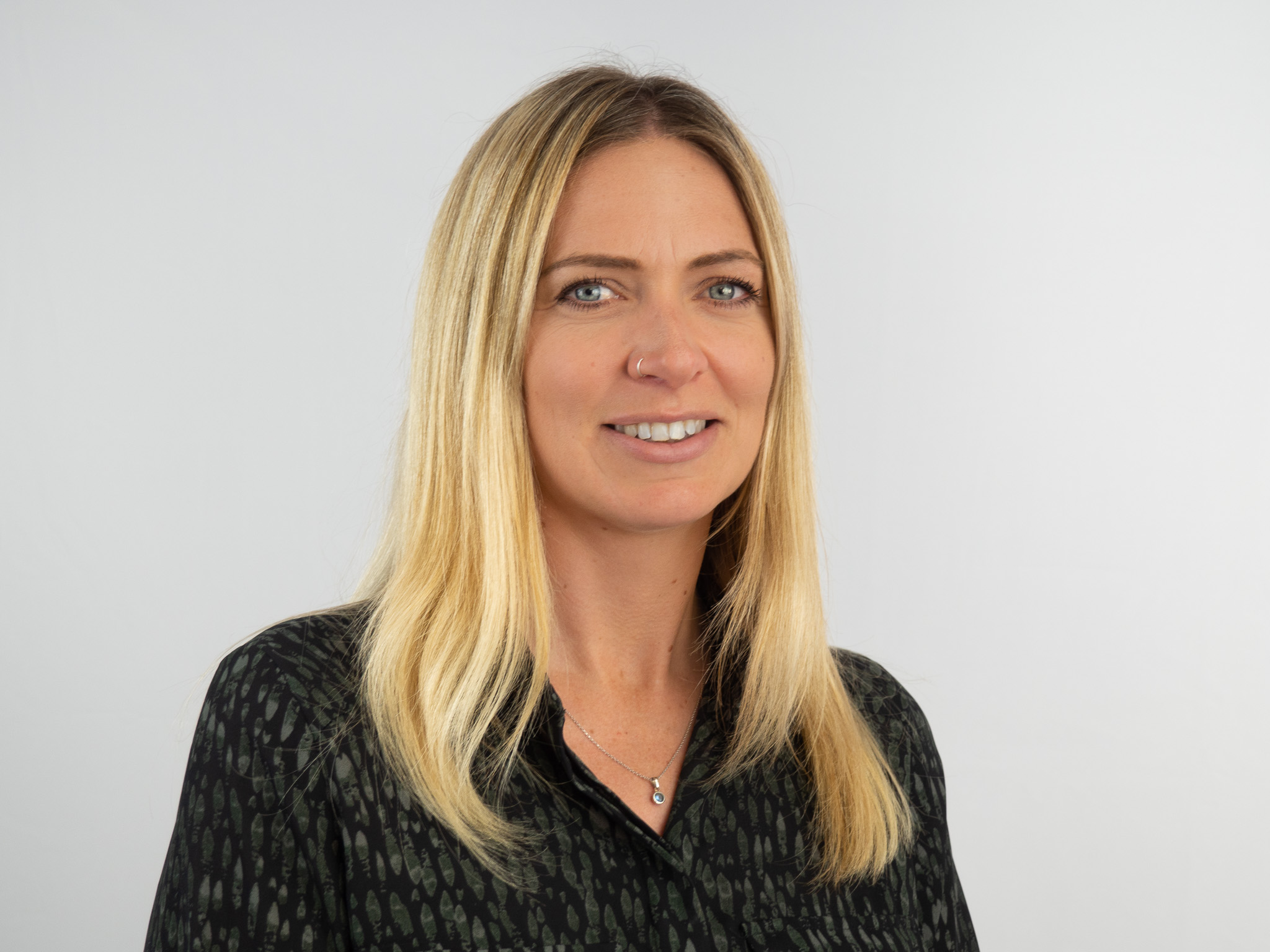 A professional headshot of a woman with shoulder-length blonde hair wearing a dark patterned blouse and a delicate necklace, smiling at the camera against a neutral gray background.