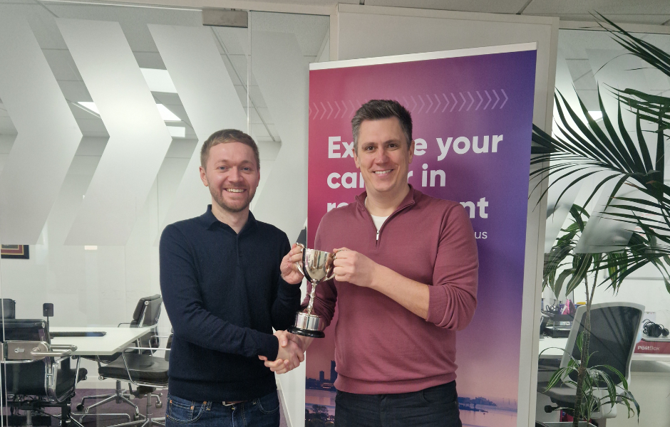 Two men smile while presenting a trophy in a modern office setting, with one man in a black shirt handing or sharing the award with another man in a burgundy quarter-zip pullover, standing in front of a purple promotional banner.