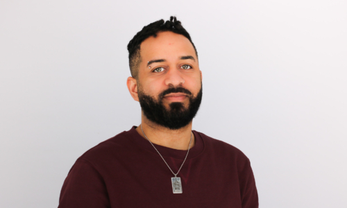 A man with dark hair and a full beard wearing a burgundy sweater and silver necklace, smiling slightly against a white background in what appears to be a professional headshot.