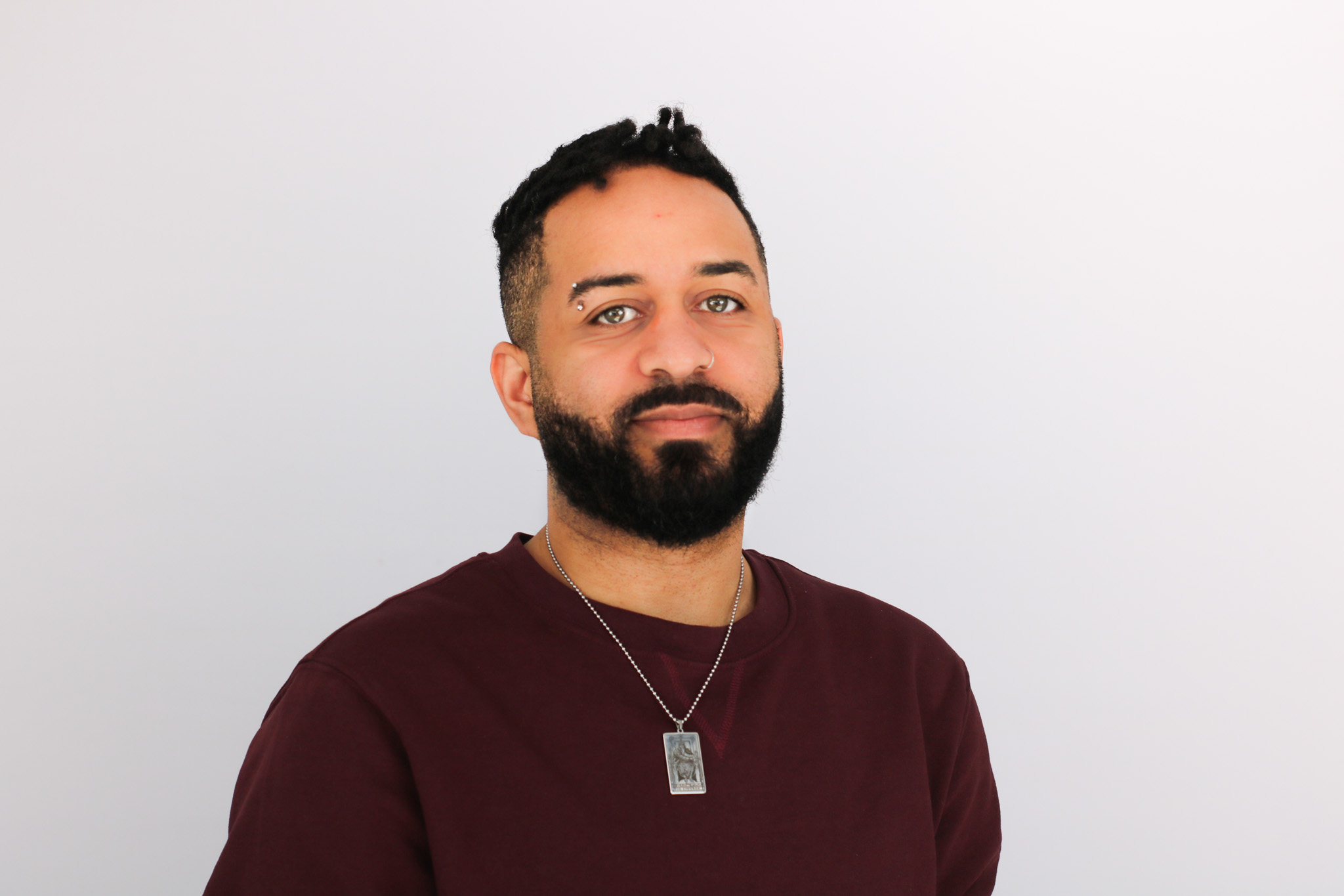 A man with dark hair and a full beard wearing a burgundy sweater and silver necklace, smiling slightly against a white background in what appears to be a professional headshot.
