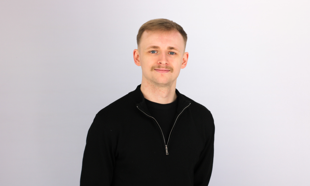 A young man with short blonde hair and a mustache smiles at the camera, wearing a black zip-up sweater against a light gray background in what appears to be a professional headshot.