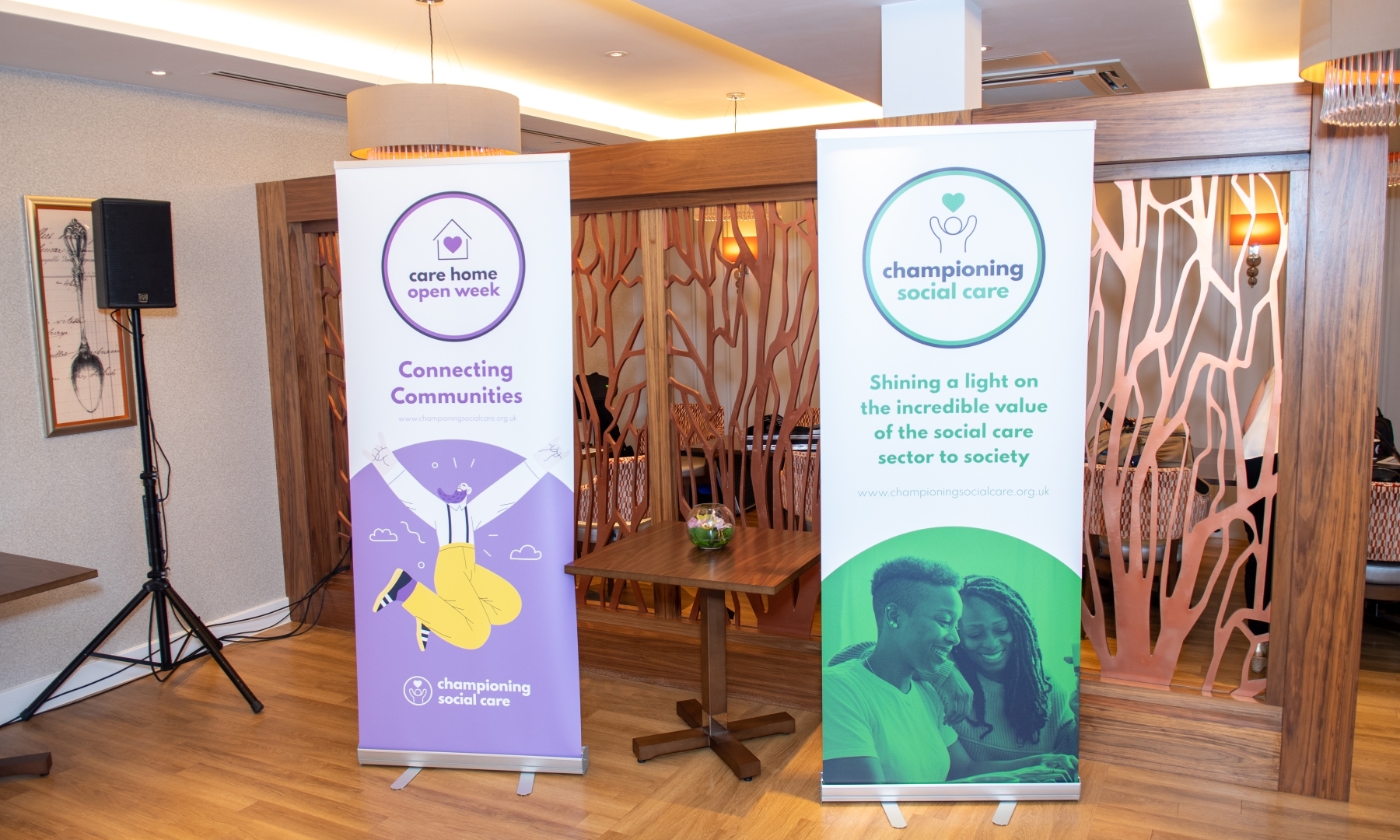 Two promotional banners stand in a modern restaurant setting, featuring "Care Home Open Week" with "Connecting Communities" messaging on a purple banner, and "Championing Social Care" highlighting the value of the social care sector on a teal and green banner with an image of two smiling women.