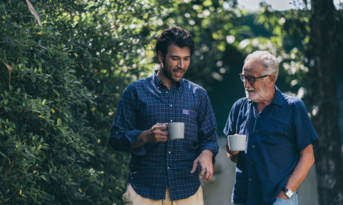 Two men of different generations walk together outdoors holding coffee mugs, with the younger man in a plaid shirt and the older man with glasses and a beard wearing a navy shirt, surrounded by lush green foliage.
