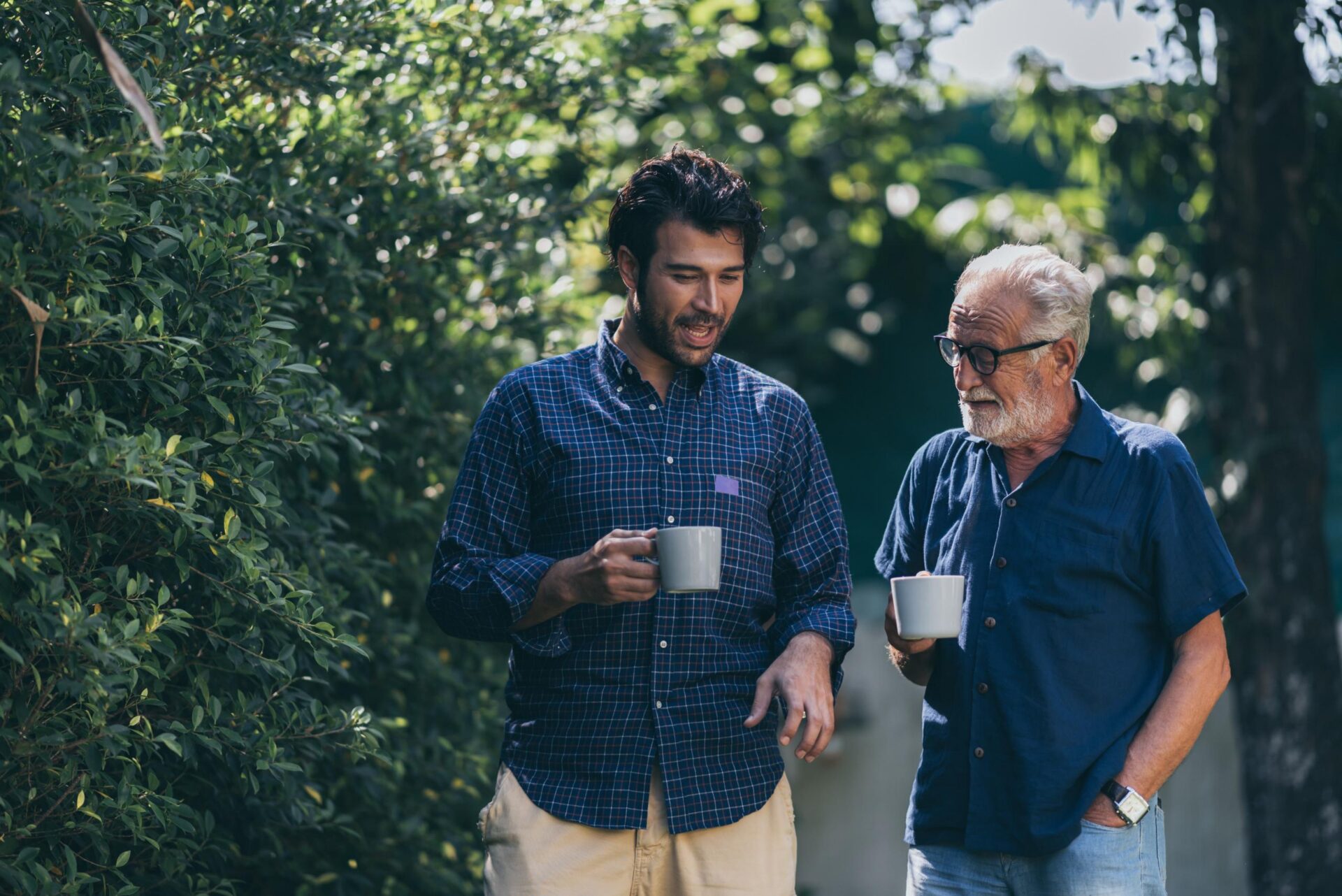 Two men of different generations walk together outdoors holding coffee mugs, with the younger man in a plaid shirt and the older man with glasses and a beard wearing a navy shirt, surrounded by lush green foliage.