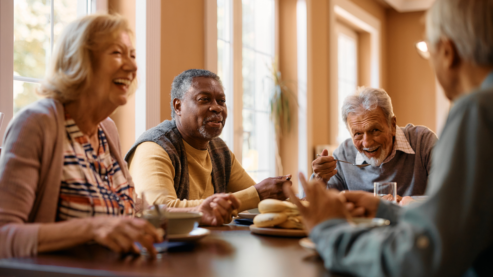 Four older adults sit around a wooden table in a bright, windowed room, laughing and enjoying a meal together. The warm, social atmosphere suggests they are sharing a pleasant dining experience, possibly in a senior living community or home setting.