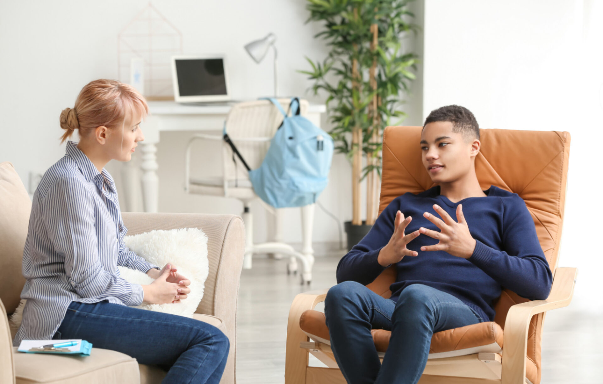 A therapist or counselor in a striped shirt sits across from a young man in a navy sweater who is gesturing with his hands while speaking during what appears to be a therapy session in a bright, modern office setting.