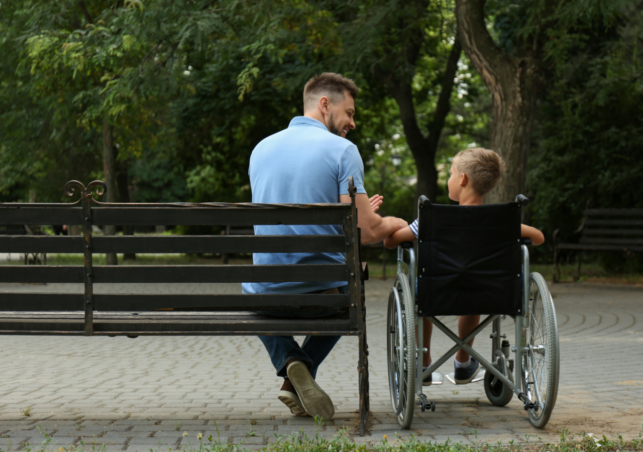 A man in a light blue shirt sits on a park bench next to a person in a wheelchair, both facing away from the camera in what appears to be a friendly conversation in a tree-lined park setting.