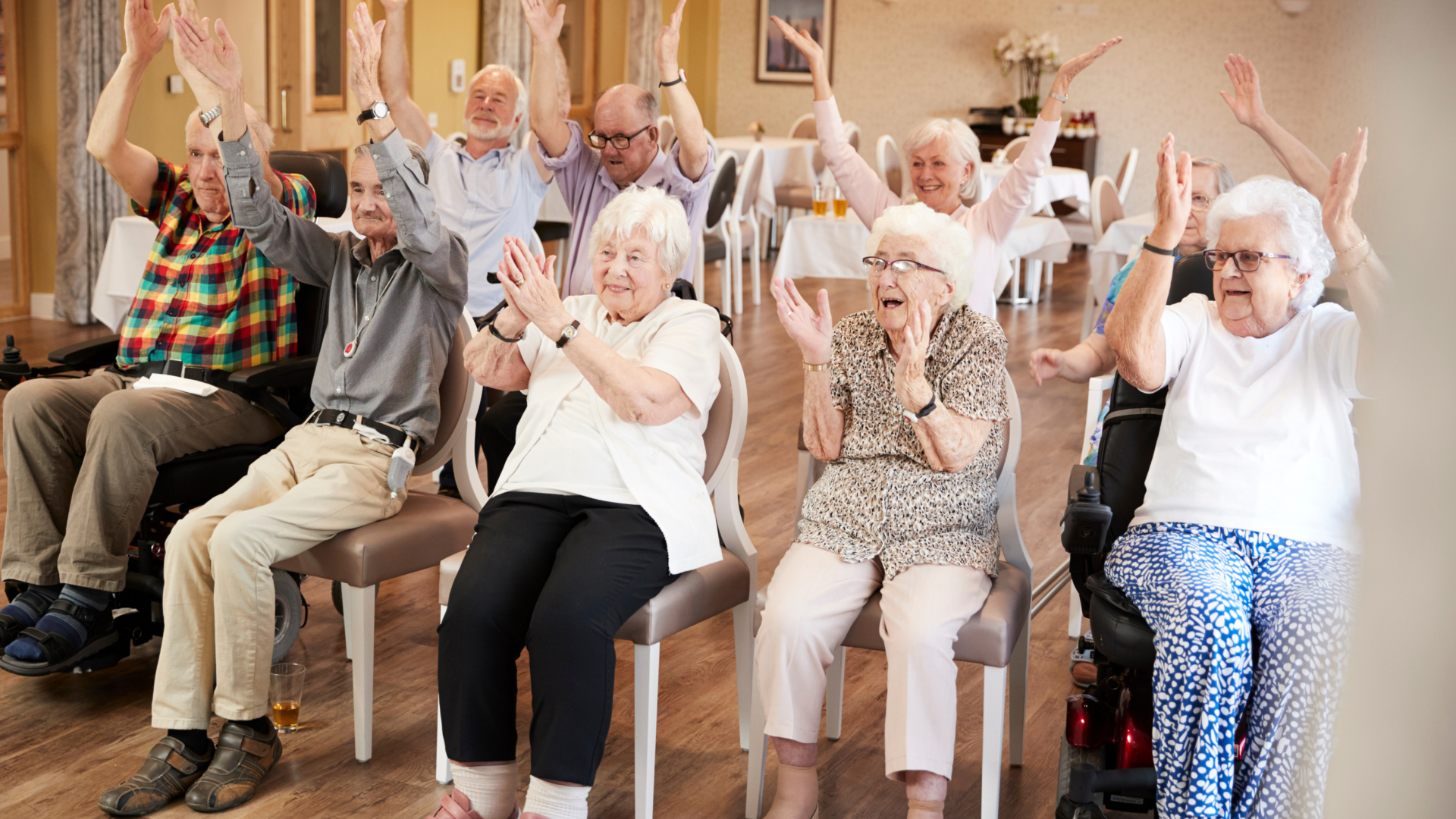 A group of elderly residents in a care home common room joyfully participate in an activity with their hands raised in the air, smiling and appearing engaged while seated in chairs and wheelchairs.