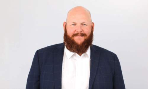 A professional headshot of a smiling bald man with a full brown beard wearing a navy blue blazer over a white dress shirt against a light gray background.
