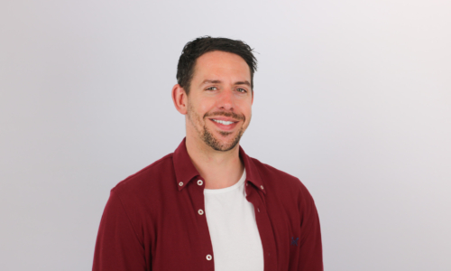 A smiling man with dark hair and facial stubble wearing a burgundy button-up shirt over a white t-shirt, photographed against a plain white background in a professional headshot style.