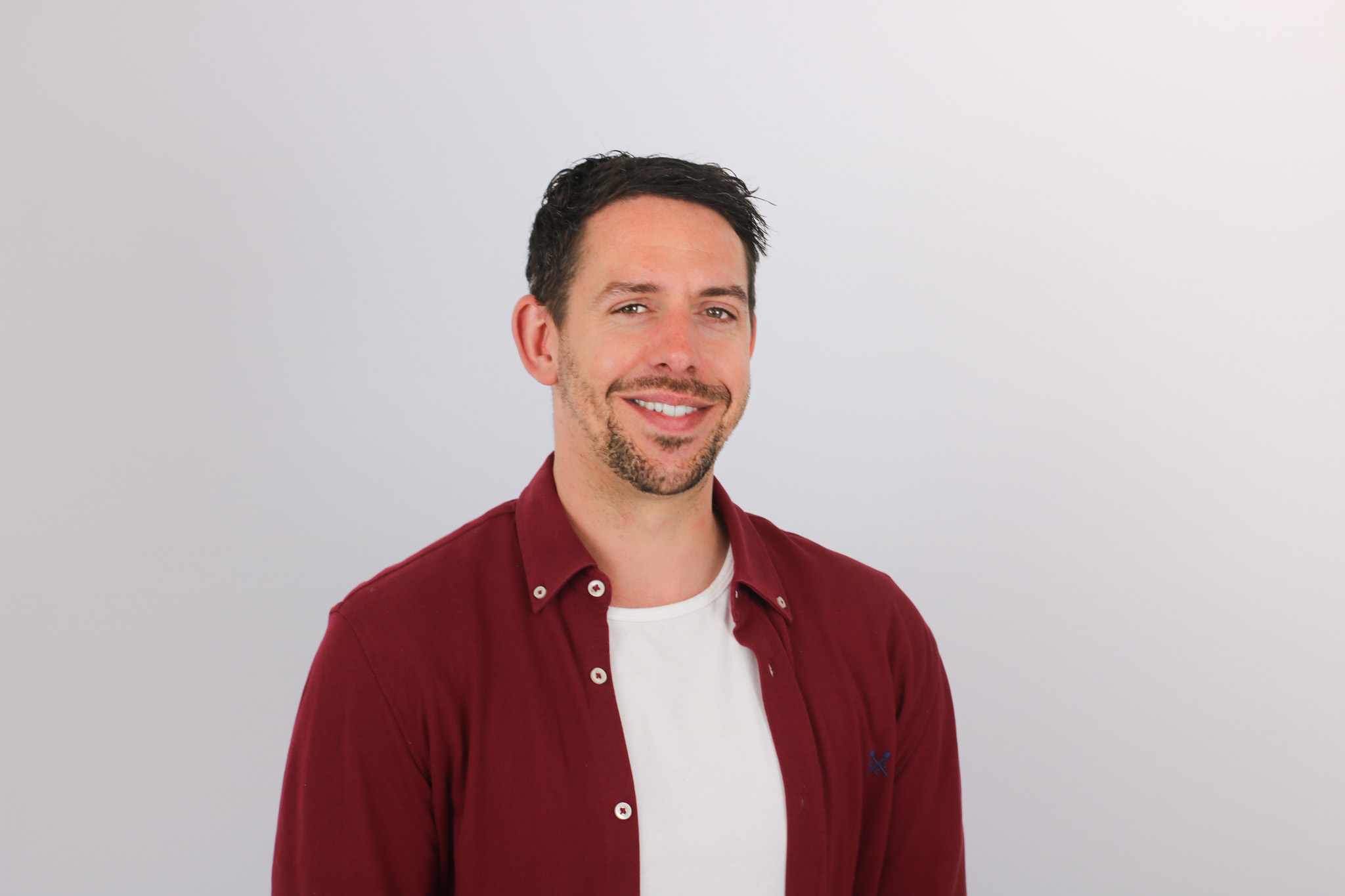A smiling man with dark hair and facial stubble wearing a burgundy button-up shirt over a white t-shirt, photographed against a plain white background in a professional headshot style.