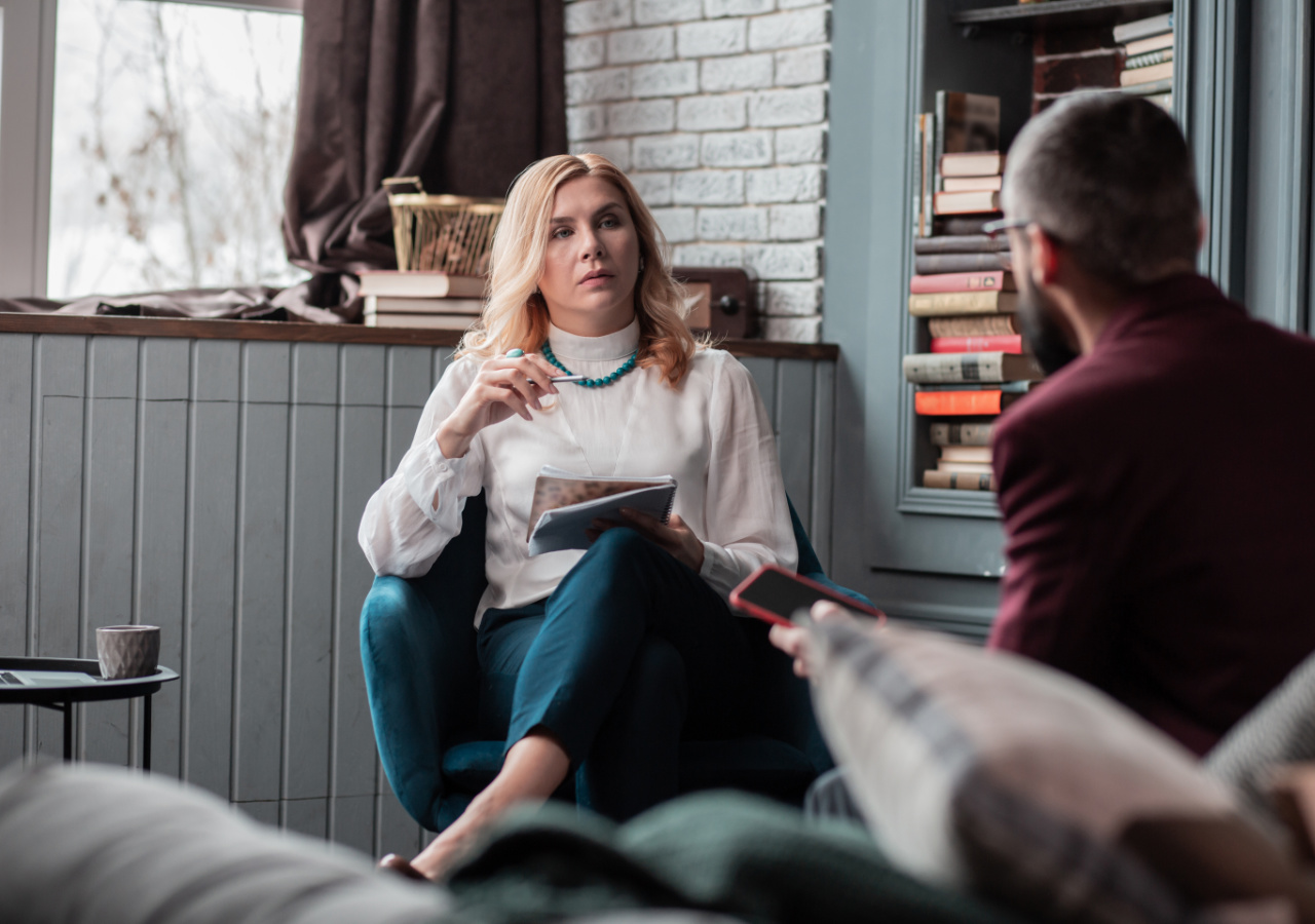 A blonde woman in a white blouse and teal pants sits in a cozy room holding a book, appearing to be in conversation with a man in a burgundy sweater who sits across from her. The setting features built-in bookshelves, white brick walls, and a window with dark curtains, creating an intimate reading or counseling session atmosphere.