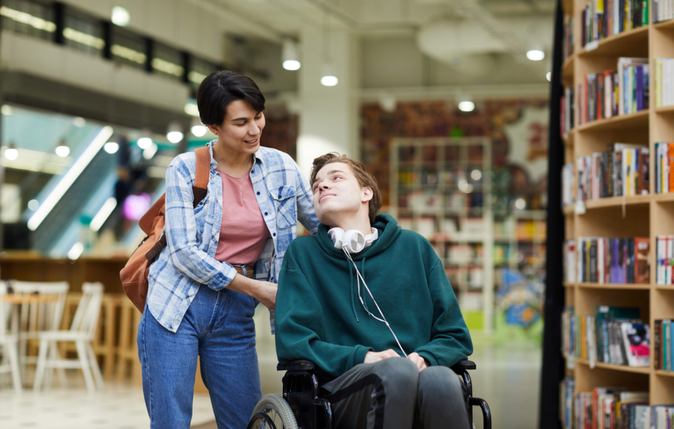 A woman stands behind a man in a wheelchair as they share a friendly moment together in a modern library with bookshelves and seating areas visible in the background. The man wears headphones and a green hoodie while the woman has a plaid shirt and carries a brown bag.
