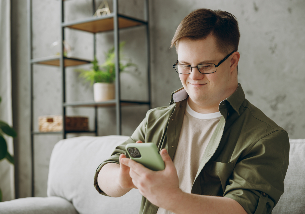 A young man with glasses and brown hair sits on a white couch, smiling while looking at his green smartphone. He's wearing a green button-up shirt over a white t-shirt in a modern living room with industrial-style shelving in the background.