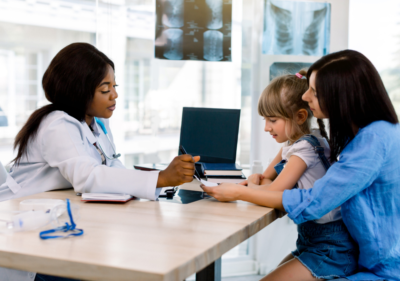 A female doctor in a white coat examines a young girl's arm while the child's mother sits beside her, with X-ray images visible on the wall behind them in a medical office setting.