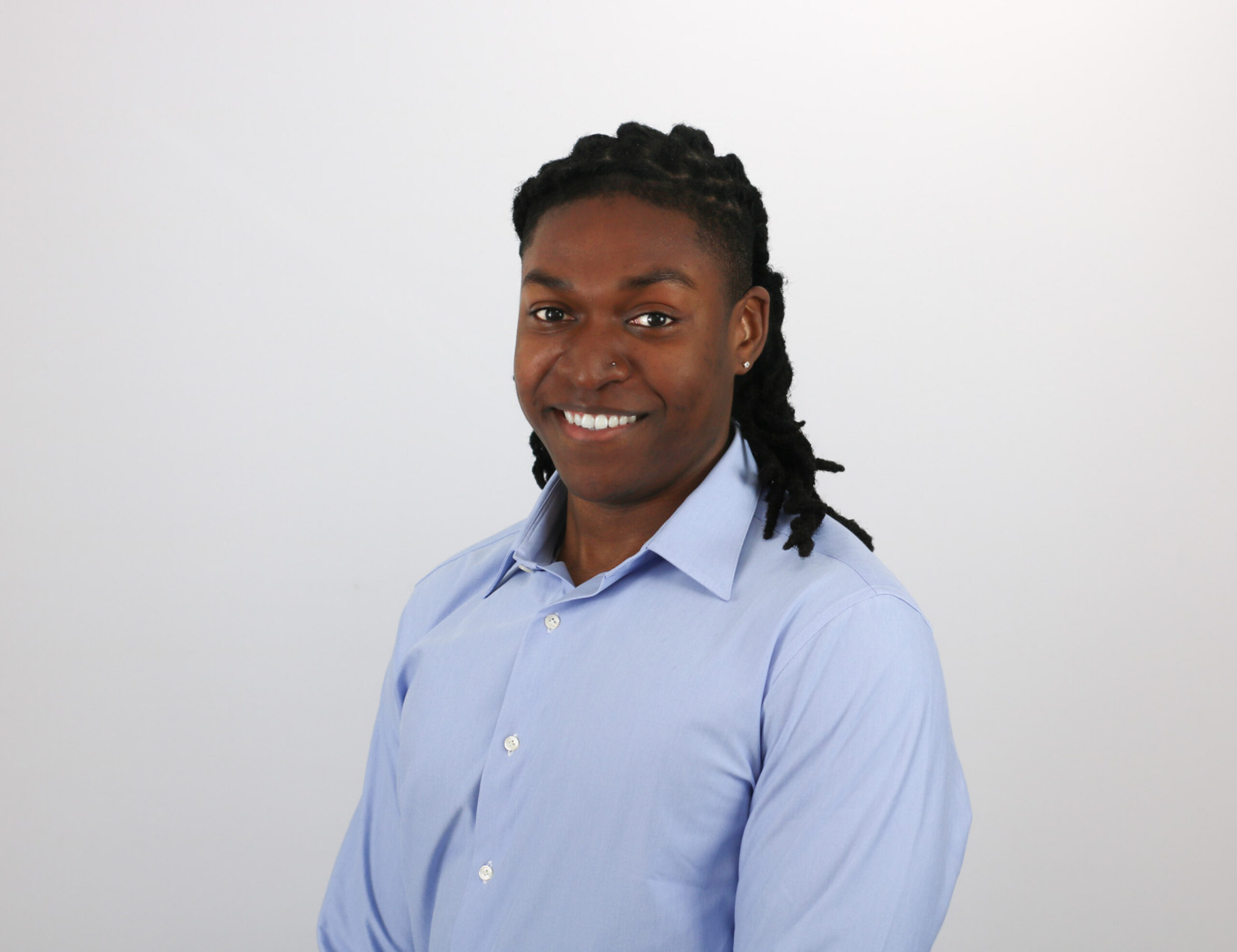 A person with dark hair styled in locs smiles warmly while wearing a light blue button-down shirt against a plain white background.