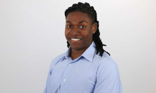 A person with dark hair styled in locs smiles warmly while wearing a light blue button-down shirt against a plain white background.