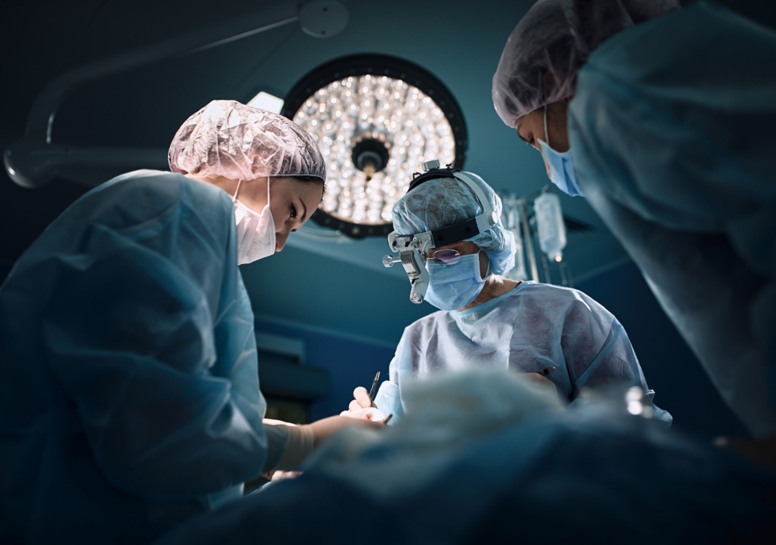 Three surgeons in surgical scrubs, masks, and head coverings perform an operation under bright surgical lights in an operating room, with one surgeon wearing a head-mounted surgical loupe for magnification.