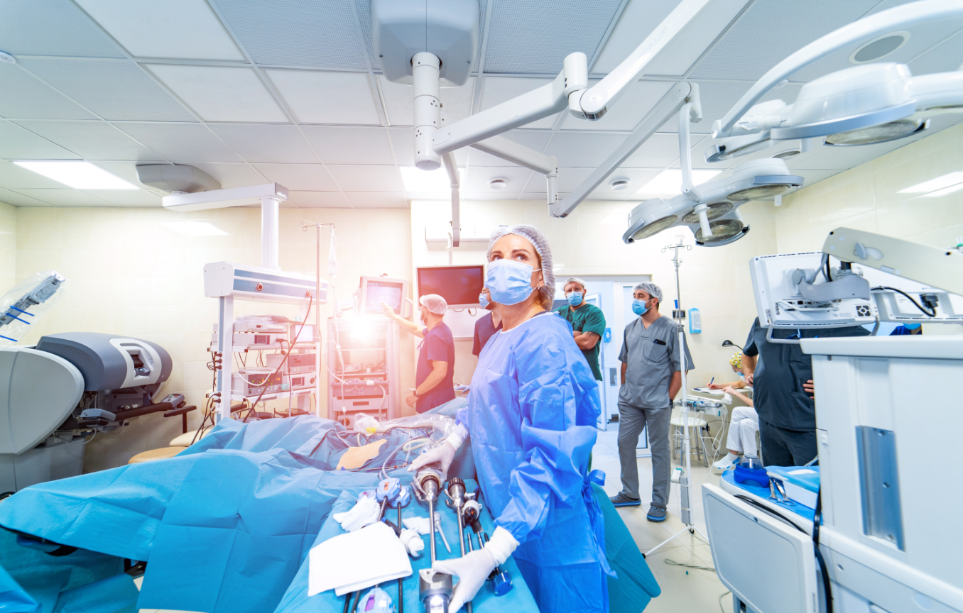 A surgeon in blue scrubs and mask performs a minimally invasive procedure using robotic surgical equipment in a modern operating room, while other medical staff observe in the background.