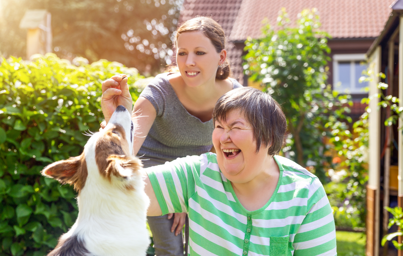 A caregiver stands behind a woman in a wheelchair wearing a green striped shirt, who is laughing joyfully while interacting with a small brown and white dog in a sunny garden setting.
