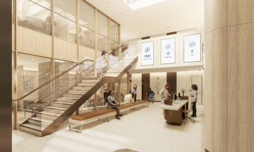A modern, bright lobby interior featuring a prominent wooden staircase with glass railings, curved wood-paneled walls, and digital displays showing statistics. Several people are visible throughout the space, including staff at a reception desk and visitors using the stairs and seating areas.