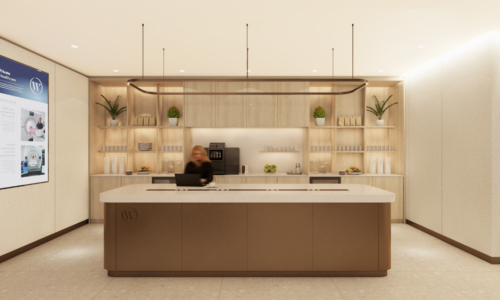 A modern commercial kitchen or coffee bar area with light wood cabinetry, white countertops, and organized shelving displaying bottles and products, with a person in dark clothing working behind the counter.