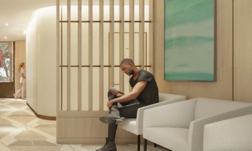 A man sits on a white modern sofa in a contemporary lobby or waiting area, looking at his phone while surrounded by wood paneling, geometric floor tiles, and a large turquoise abstract painting on the wall.