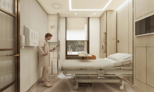A healthcare worker in scrubs stands beside a hospital bed in a modern, well-lit patient room with beige cabinetry, medical equipment, and a window with natural lighting.