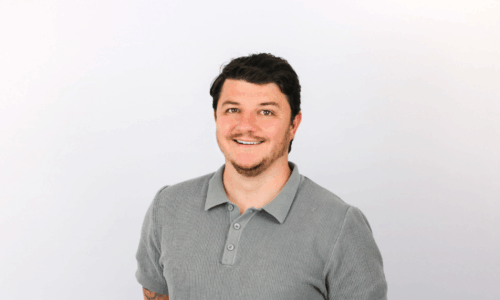A smiling man with dark hair and facial stubble wearing a gray ribbed polo shirt against a white background.
