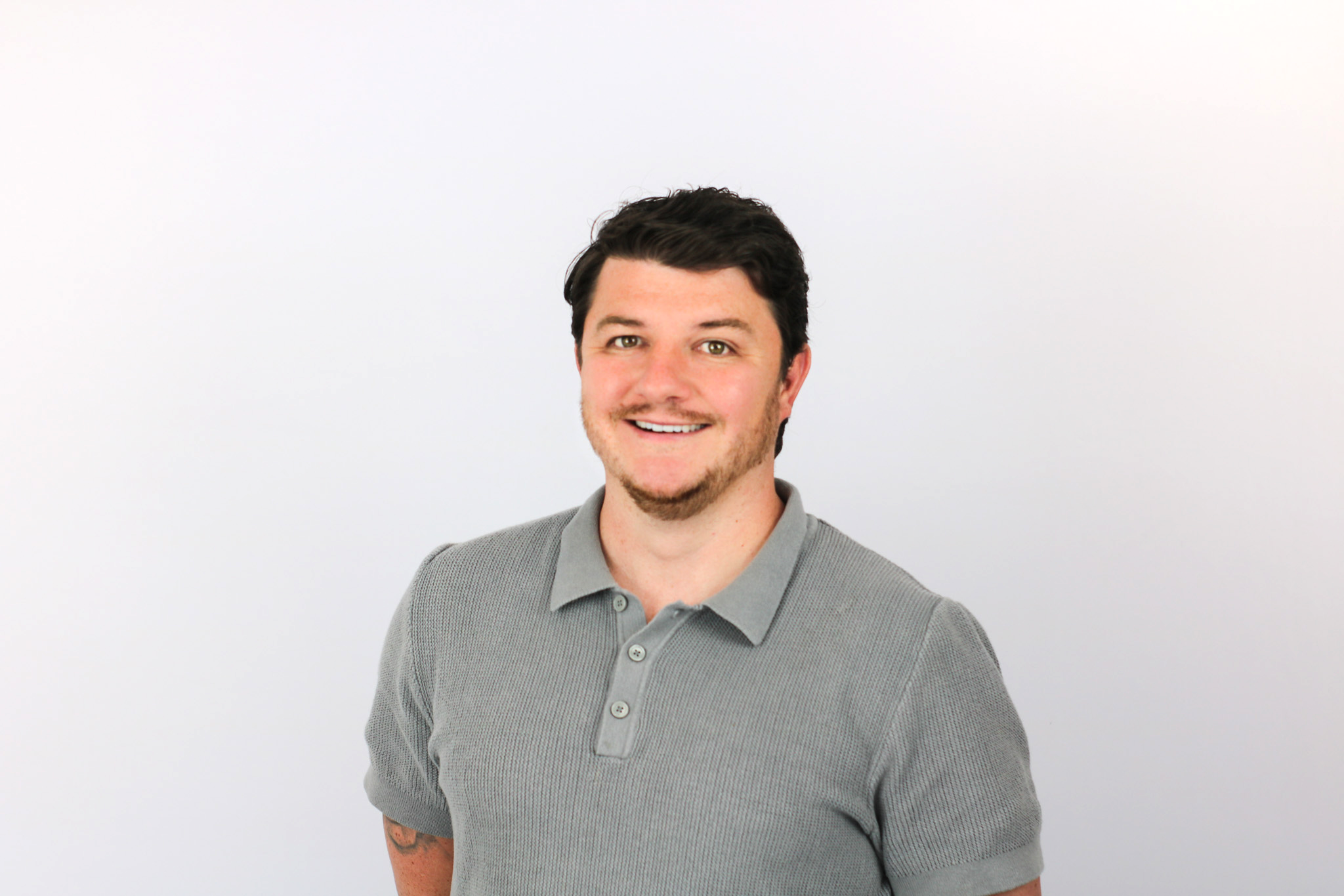 A smiling man with dark hair and facial stubble wearing a gray ribbed polo shirt against a white background.