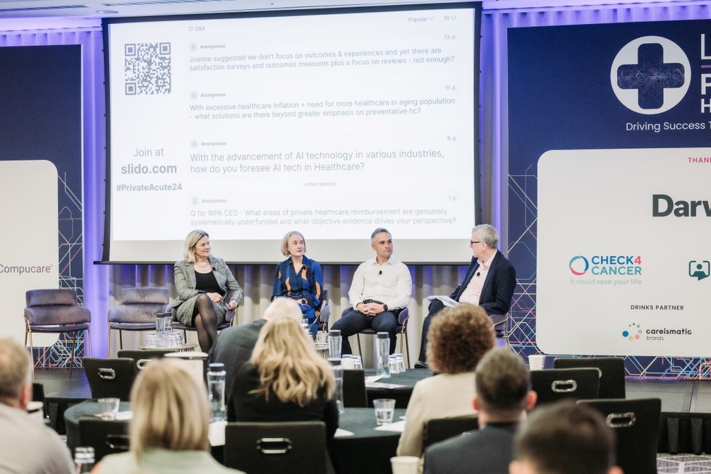 A panel of four healthcare professionals sits on stage at a conference discussing private healthcare topics, with a large presentation screen behind them displaying questions about AI technology in healthcare and a QR code for audience participation via slido.com.