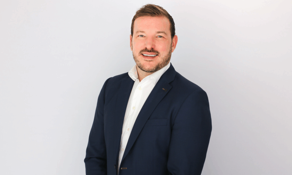 A smiling man with brown hair and facial hair wearing a dark navy blazer over a white shirt, photographed against a light gray background in a professional headshot style.
